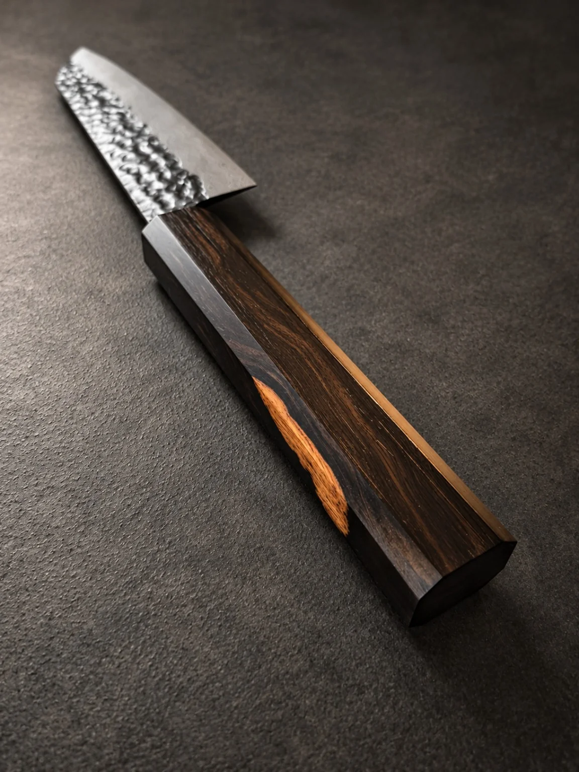 Japanese-style kitchen knife with a dark figured wooden handle and a dramatic sapwood streak, photographed diagonally against charcoal stone.